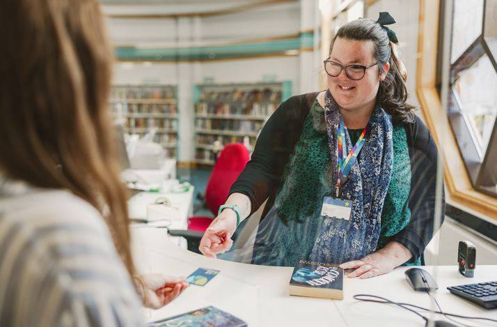 An image of a smiling library worker behind a desk taking a library card from a customer. She is smiling and happy.