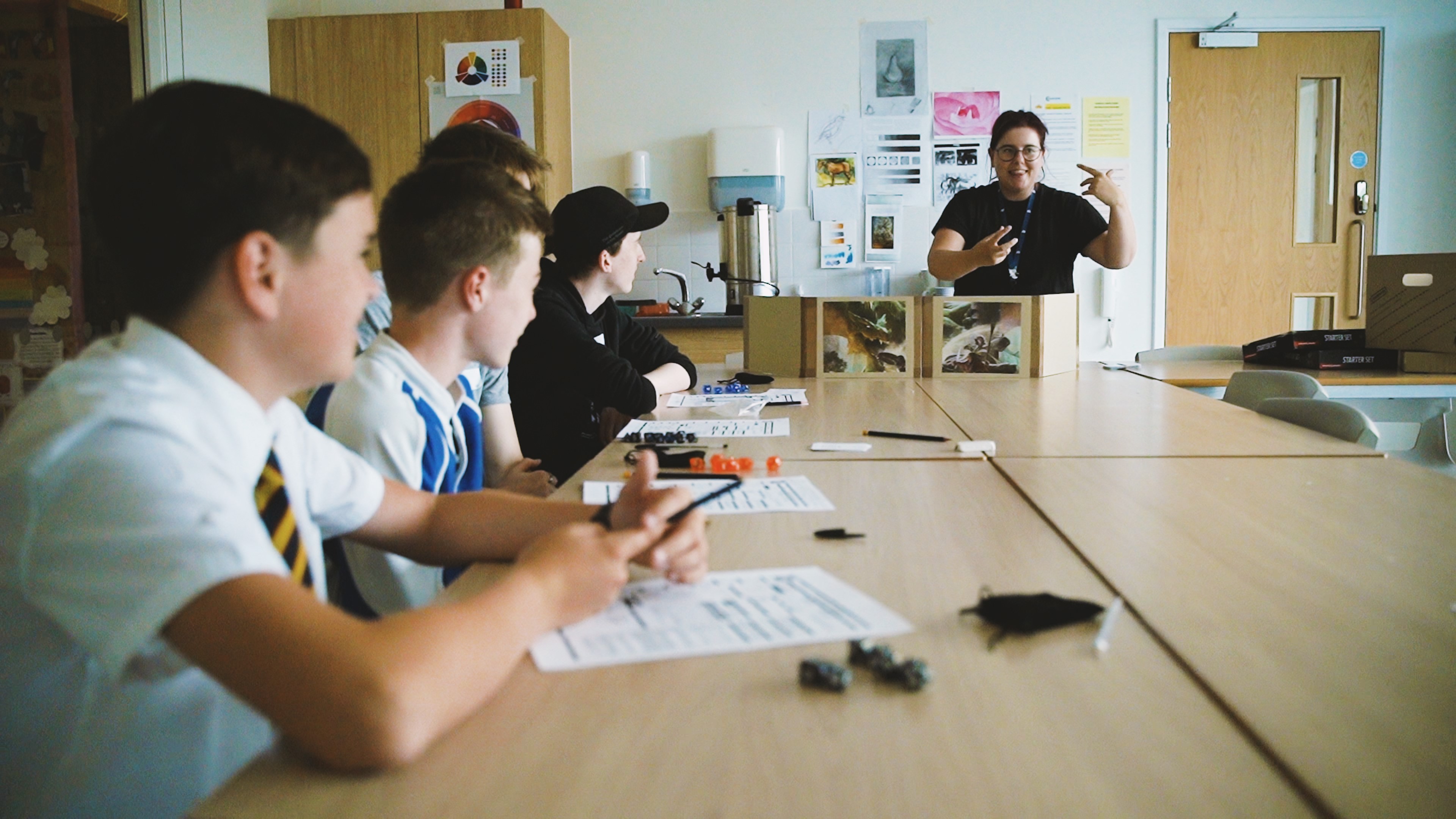 An image of school children sat around a table listening to a teacher.