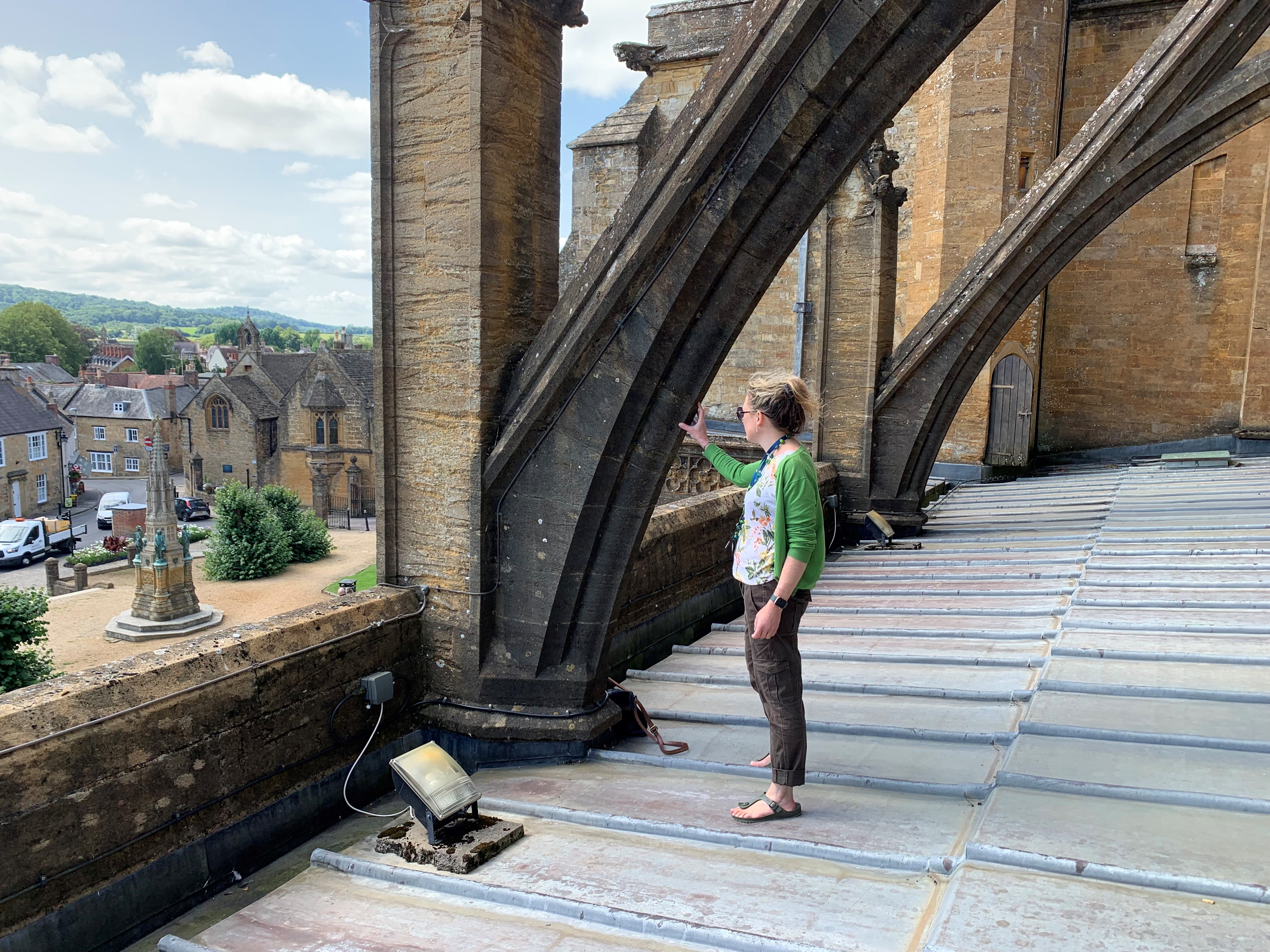 A woman stands on top of Sherborne Abbey roof, touching the stone and looking at the landscape of house roofs below.