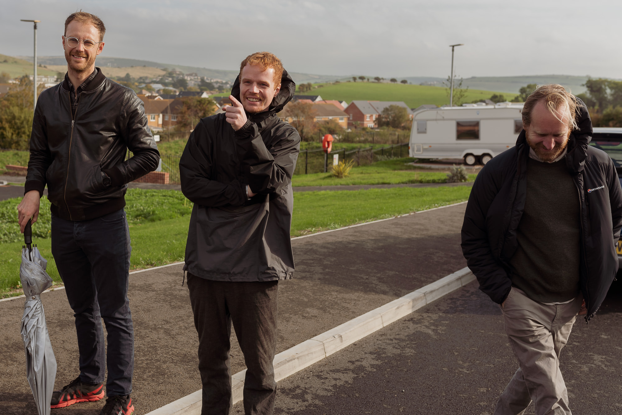 Three men on a site visit. One has an umbrella, one is pointing. There are hills and a caravan in the background.
