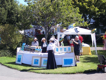 picture of a stall at a fete which is selling framed prints. There is a lady in Victorian clothing at the front of the stall, and other people in costume behind it. A man is taking a picture of them.