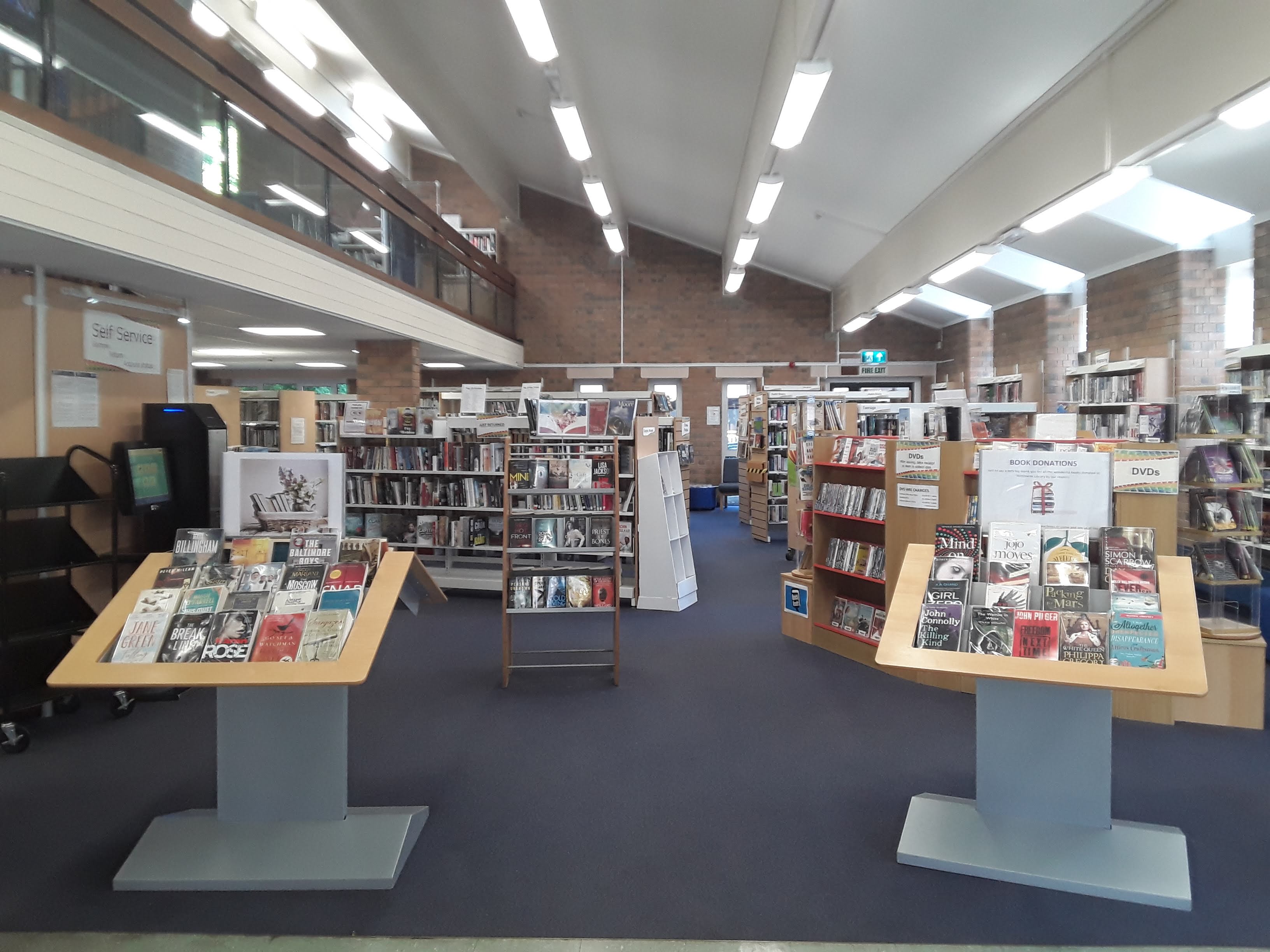 Picture of interior of the ground floor of Wimborne library. There is a balcony on the left of the picture, and light from windows on the right. You can see the back wall behind lots of book cases and display stands.