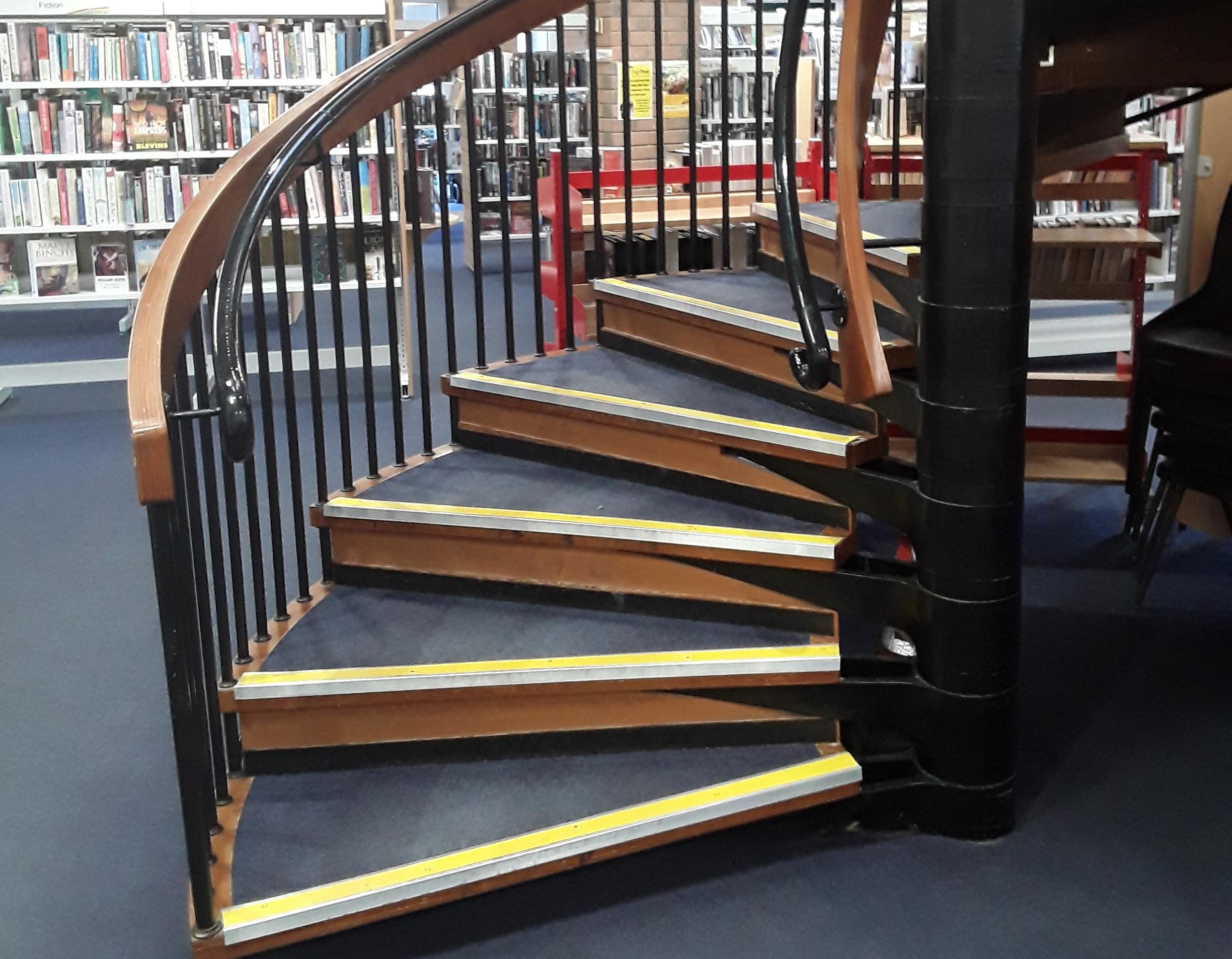 The bottom of the spiral staircase in the library. The steps have handrails either side and high visibility yellow stripes on each riser.
