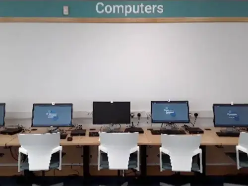This picture shows a long desk with five computer screens and keyboards and four chairs in front.