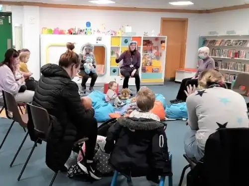 This picture shows a group of adults and children attending a rhythm time session.  The room is full of chairs and there are soft cushions in the centre for the children.