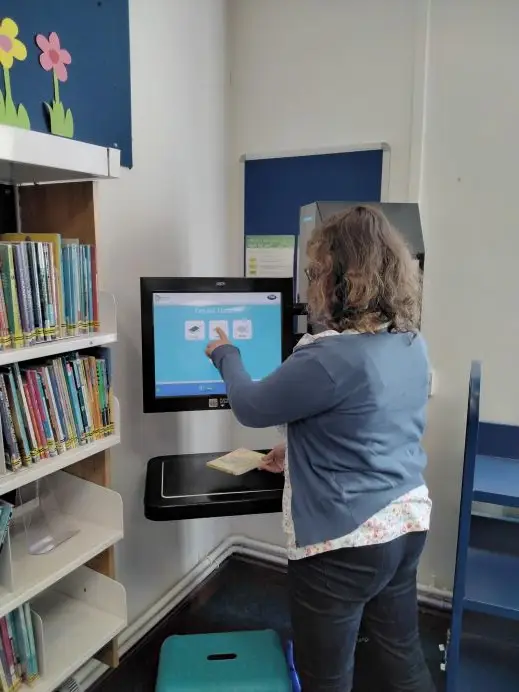 This is a picture of a library assistant standing in front of a library self service machine, explaining how it is used.