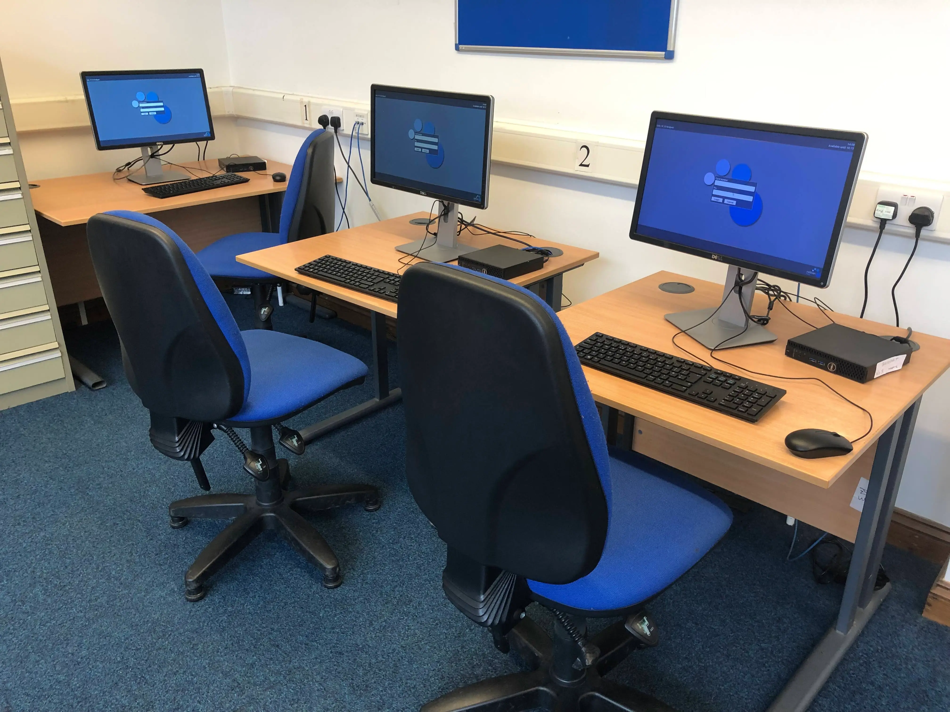 This is a picture of three desks with computer screens and keyboards on each.  There is a blue chair in front of each desk.