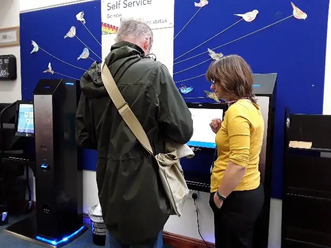 This is a picture of two people, a library assistant and customer and the library assistant is helping the customer at the self service machine.