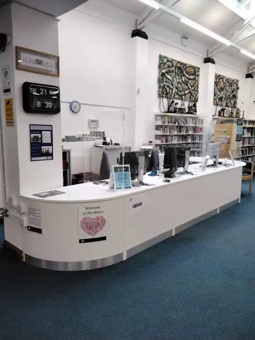 This is a picture of a white front desk with three computer screens and keyboards behind plastic screen on desk.  There is a clock on the back wall.