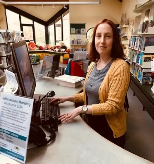 This is a picture of a library assistant sitting behind a desk with a computer screen and keyboard in front.  They are looking at the camera.