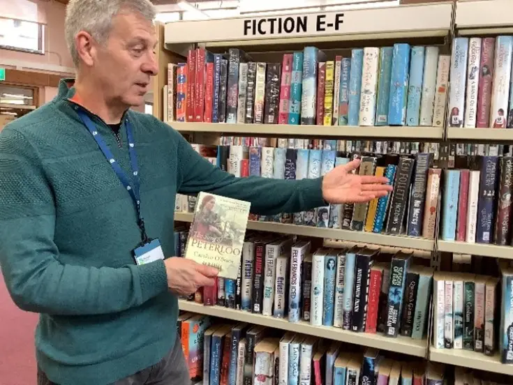 This is a picture of a library assistant holding a book and pointing at the books on a shelf.