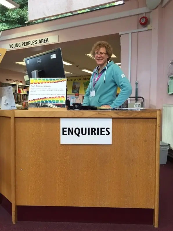 This is a picture of the enquiry desk with a library assistant standing behind it smiling at the camera.