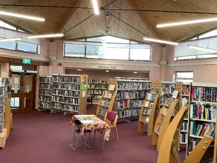This is a picture of a large room full of shelving with books on.  There is a table with two chairs to the front of the picture.
