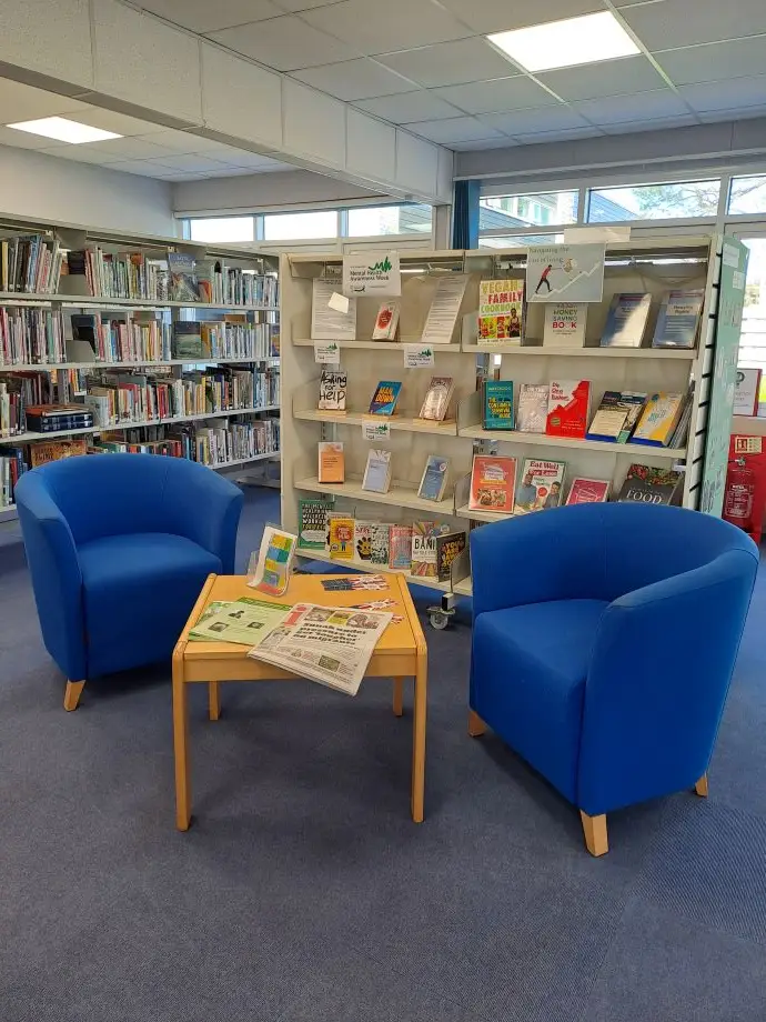 This is a picture of a small square table and two blue chairs with rows of shelving with books behind.