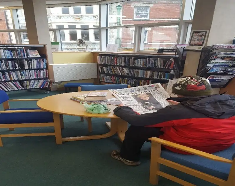This is the picture of a person reading a newspaper at a round table with two other chairs.  There are book shelves full of books behind the table.