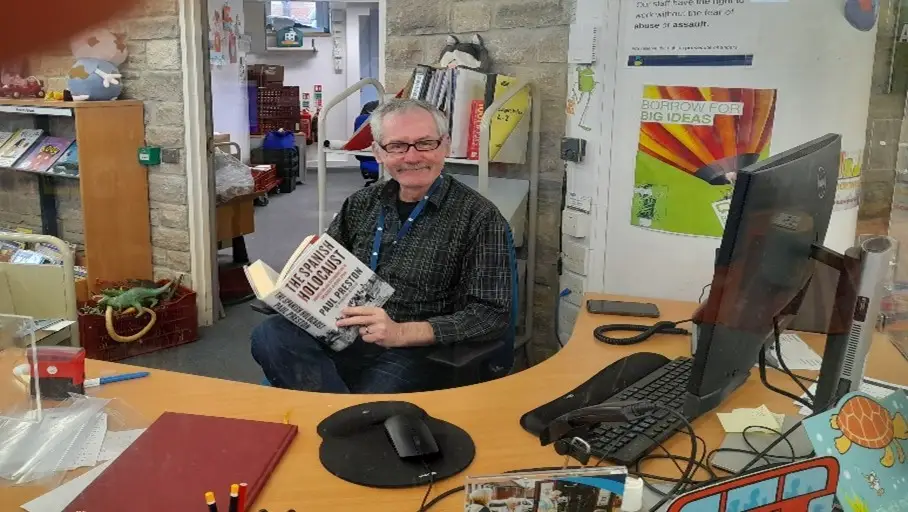 This is a picture of a library assistant behind a desk holding a book and smiling at the camera.