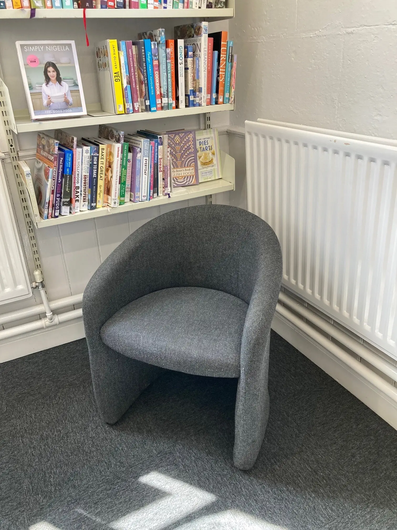This is a picture of a grey chair in front of shelving unit with books on.  There is a radiator to the right.
