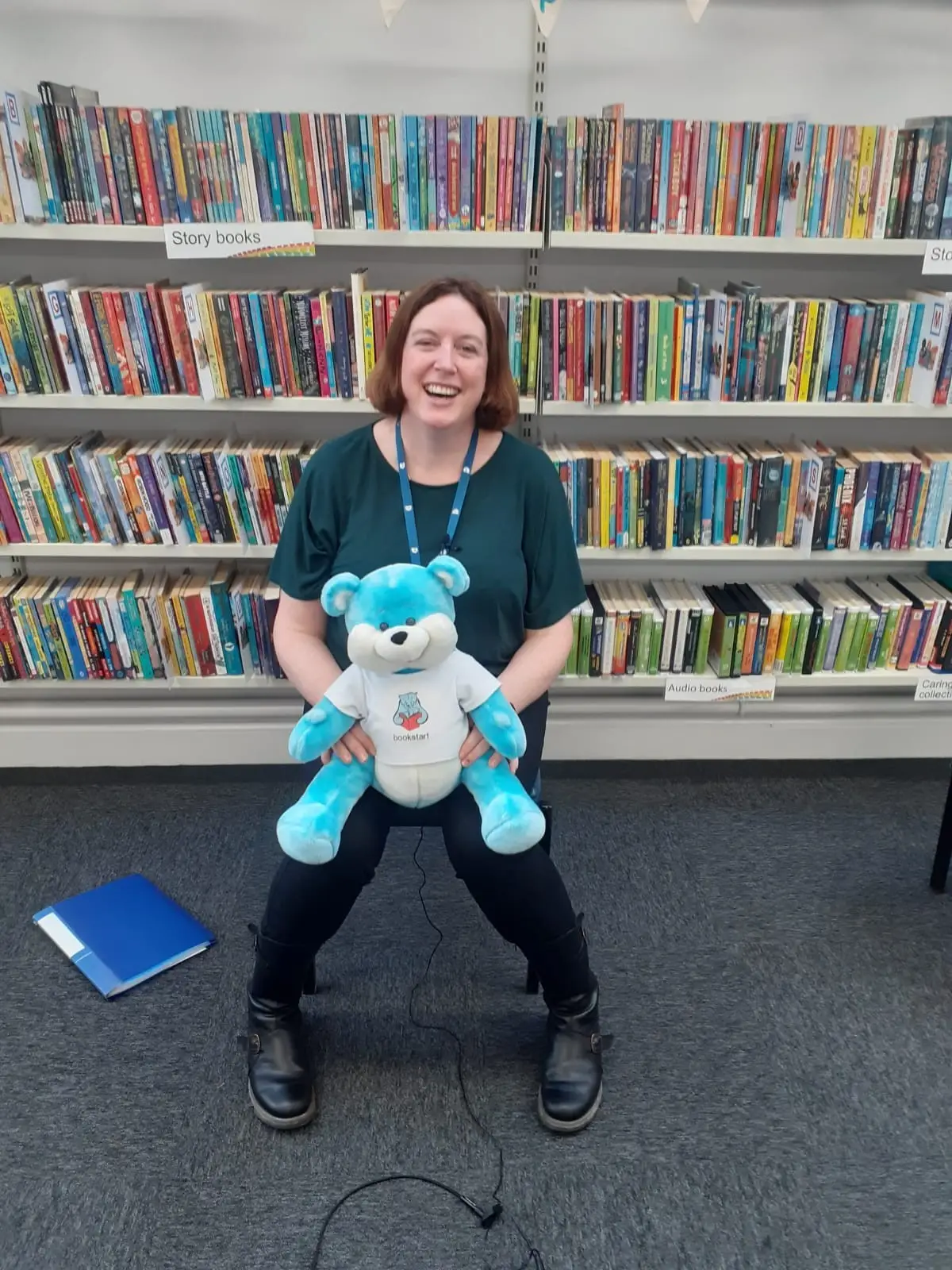 This is a picture of a library assistant holding a blue Bookstart Bear sitting down and smiling at the camera,