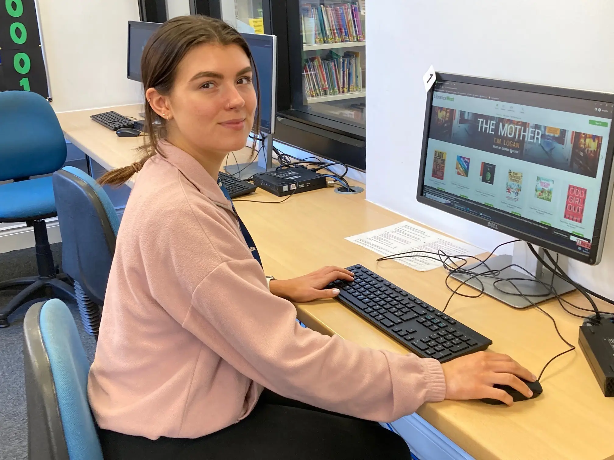 This is a picture of a library assistant sitting in front of a computer screen and keyboard, turning around smiling at the camera.