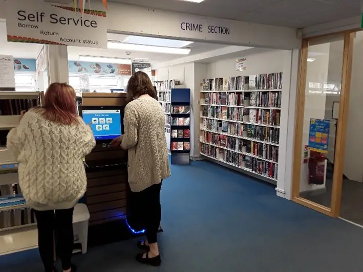 This is a picture of a library assistant helping a customer use the self service machine to take out books.