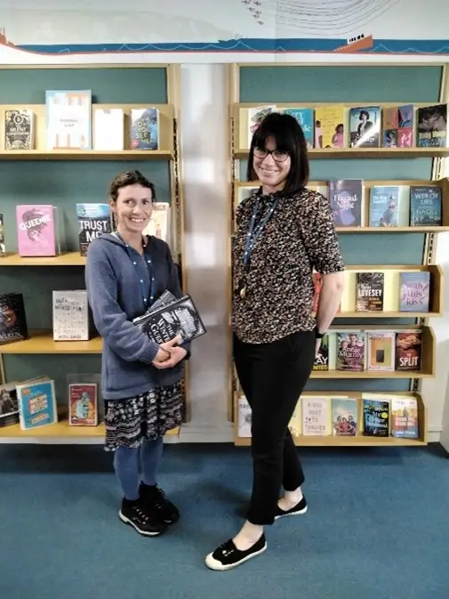 This is a picture of two library assistants standing and smiling at the camera in front of displays of books.