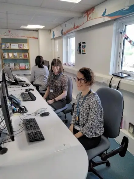 This is a picture of the front desk with three library assistants sitting on chairs behind desk.  They have computer screens and keyboards in front of them.