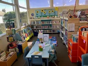 This is a picture of the children's area of the library with a small table surrounded by chairs.  There is a bookstart bear sitting on one of the chairs.  The table is surrounded by shelves of books.