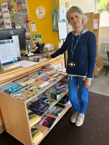This is a picture of a library assistant standing in front of a desk with a computer screen on.  There is shelving with leaflets to the front of picture.