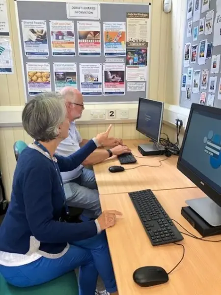 This is a picture of a library assistant helping a person to log in to the computer.  Both people are sitting in front of computer screens.