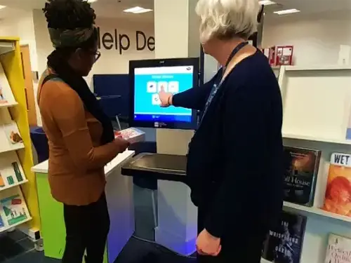 This picture shows a member of staff showing a customer how to use the self service machine.