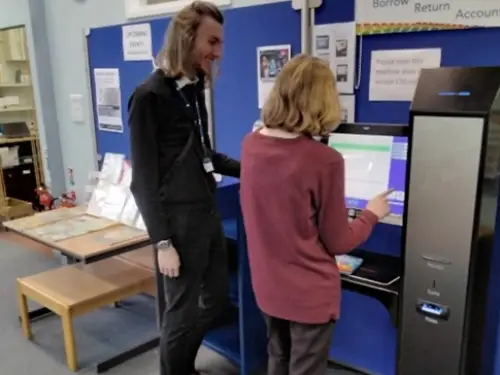 This is an image of a library assistant helping a member of the public check out a book using the self service machine.