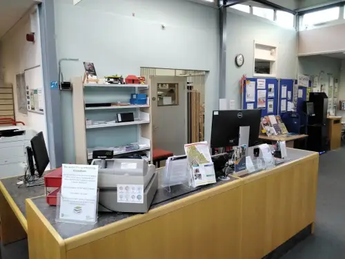 This is an image of the front desk in Sherborne Library. There is a computer screen, a cash machine and leaflets on the desk.