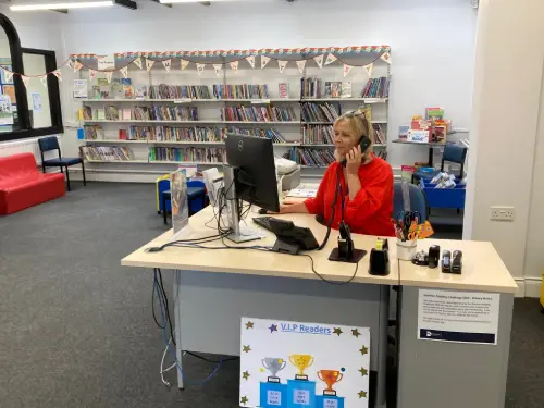 There is a library assistant sitting behind the front desk taking a call while looking at the computer screen.