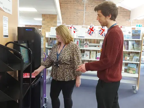 This is an image of a library assistant showing a member of the public how to check out a book at the self service machine.