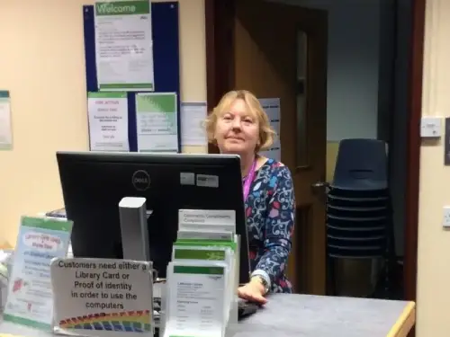 This is an image of a library assistant standing behind a computer screen at a desk looking at the camera.