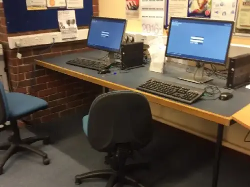 This is an image of a long desk with two computer screens and keyboards and chairs in front of desks.