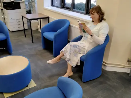 This is an image of a person reading a book in a blue chair.  There are four blue chairs in the image and a small round table.