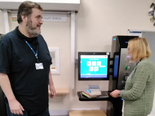 This is an image of a library assistant helping a customer use the self service machine.