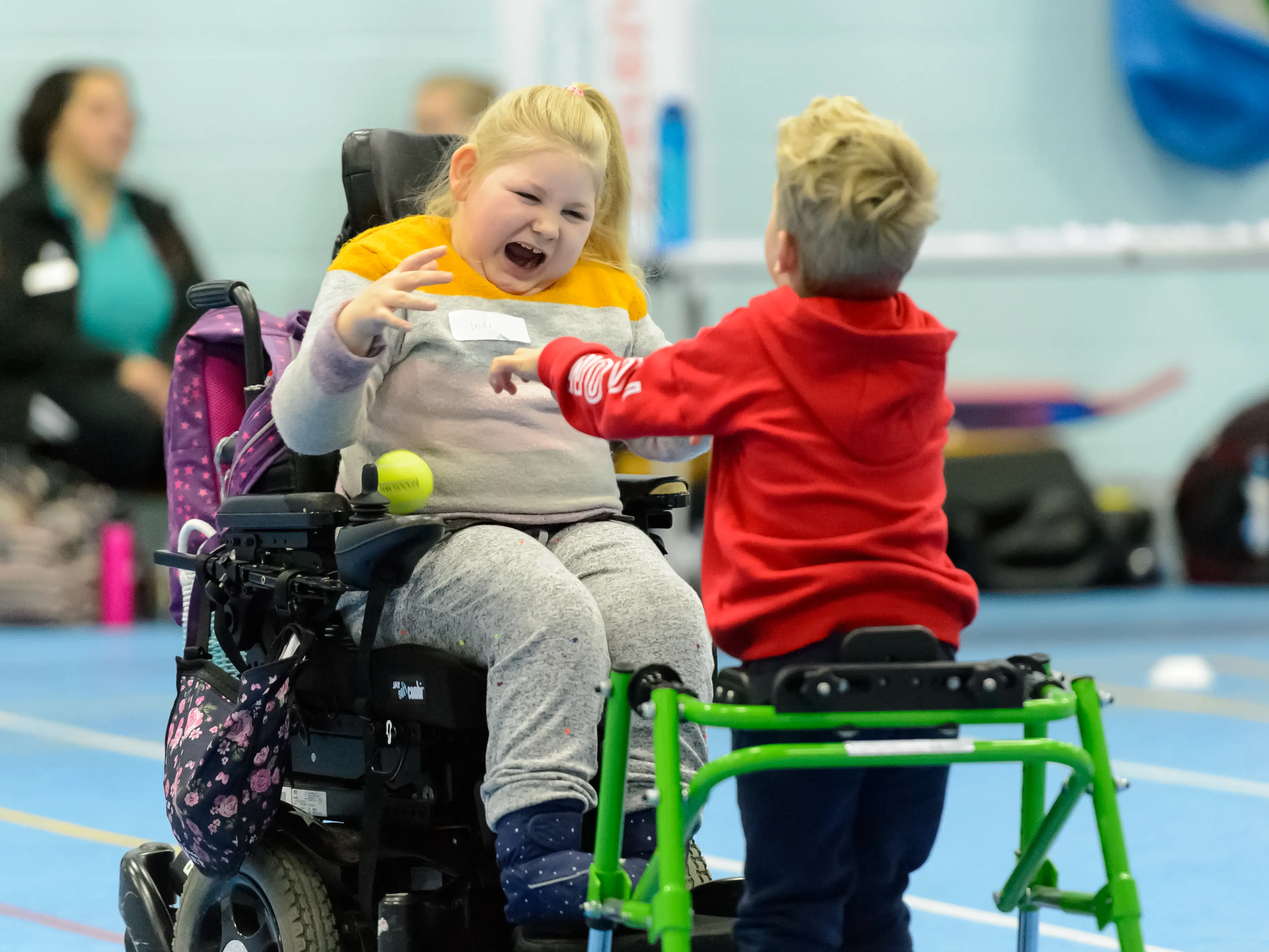 Young girl in a powered wheelchair laughing and young boy using a walking frame throwing a tennis ball to her