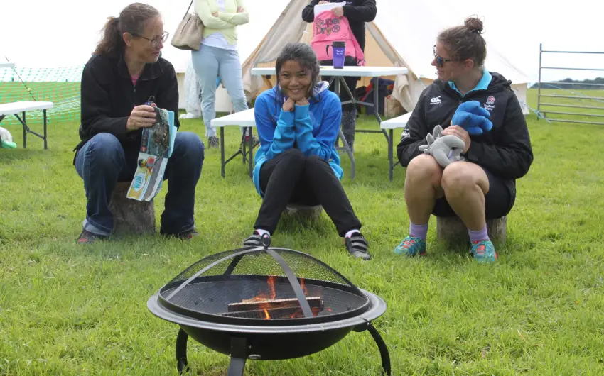 two Artz+ Sportz+ staff sit on tree stumps either side of a smiling girl all around a firepit