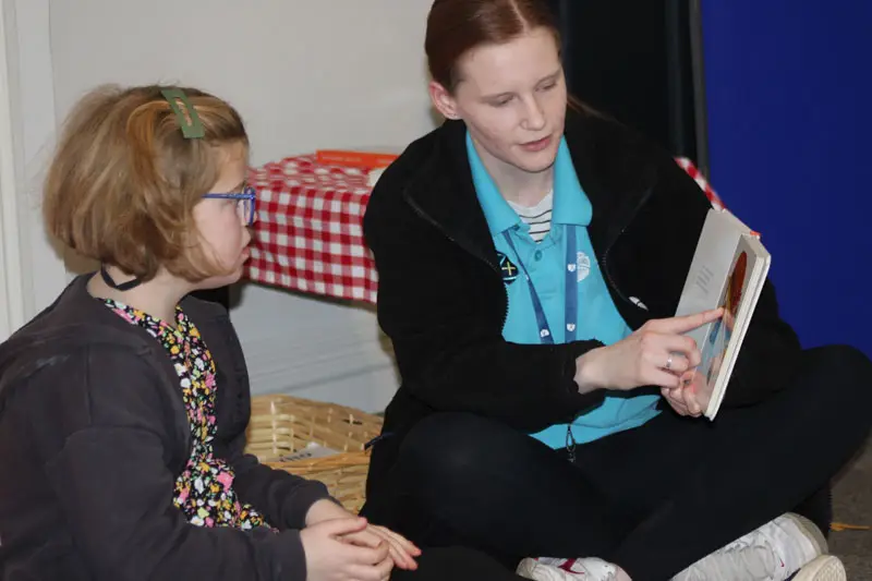 Artz+ Sportz+ female staff member reading to girl in glasses