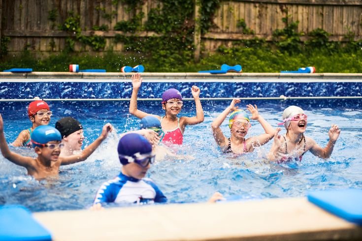 Children at Broadmayne First School splashing in an outdoor pool, wearing swim caps and goggles.