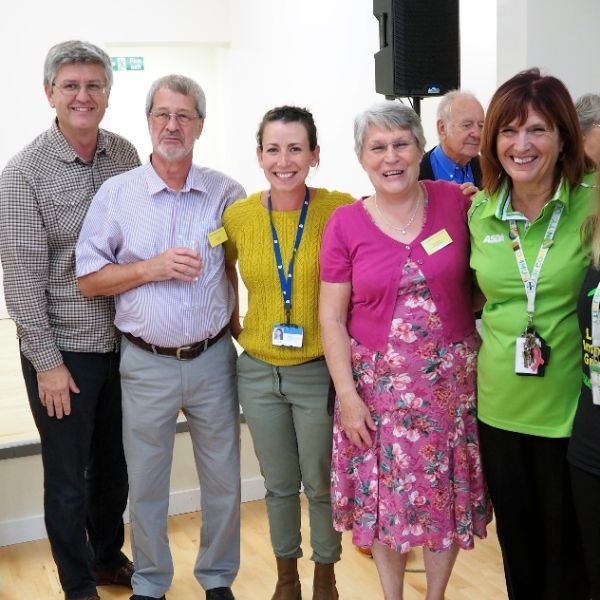 Group of six people standing together at an event in Charlton Marshall, some wearing name tags and lanyards, with a stage and speaker in the background.