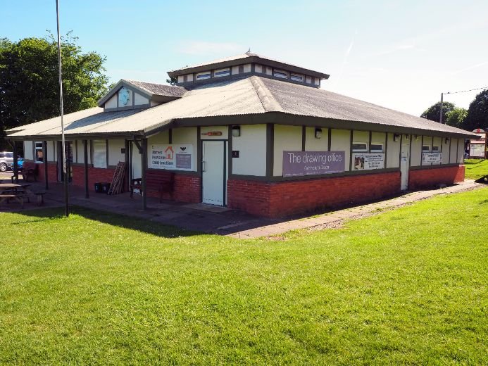 Exterior of the Dorchester Town Council Cricket Pavilion, a single-story building with a red brick base, light-colored walls, and dark trim. The building is surrounded by grass and trees under a clear, sunny sky.