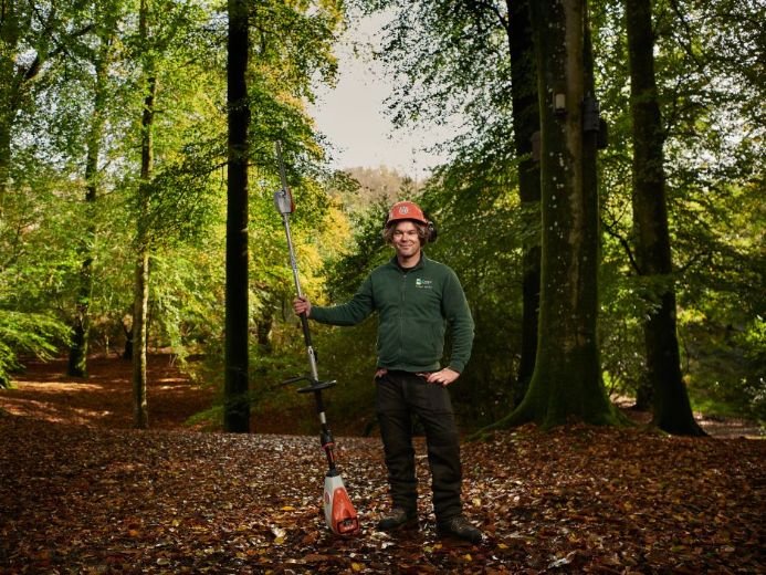 A person wearing safety gear, including an orange helmet and ear protection, stands in a forested area holding a long-handled tool with an orange and white base, surrounded by tall trees and fallen leaves.