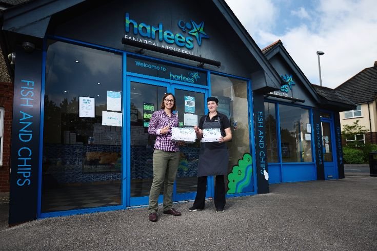 Two people holding certificates outside Harlees Fish and Chips restaurant in front of signage reading 'Fanatical About Fish'.