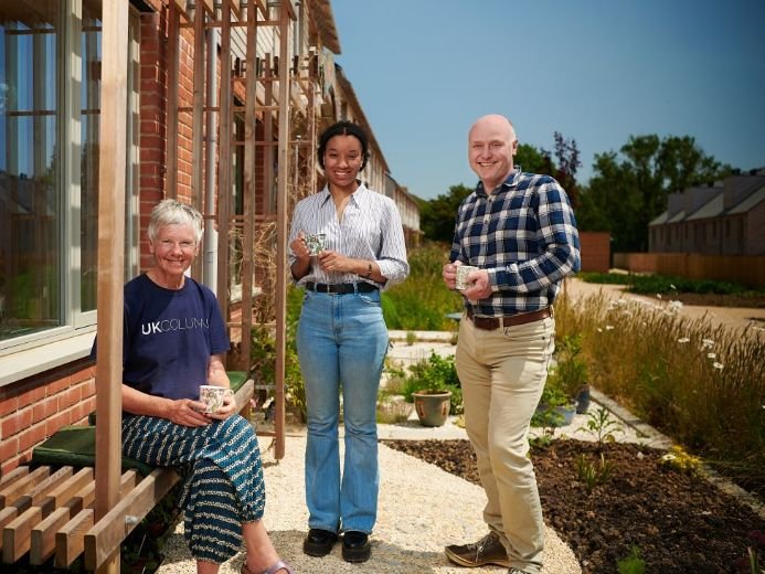Three people outside a row of houses, each holding a mug. One is seated on a bench, while the other two stand nearby, with gardens lining the pathway behind them.
