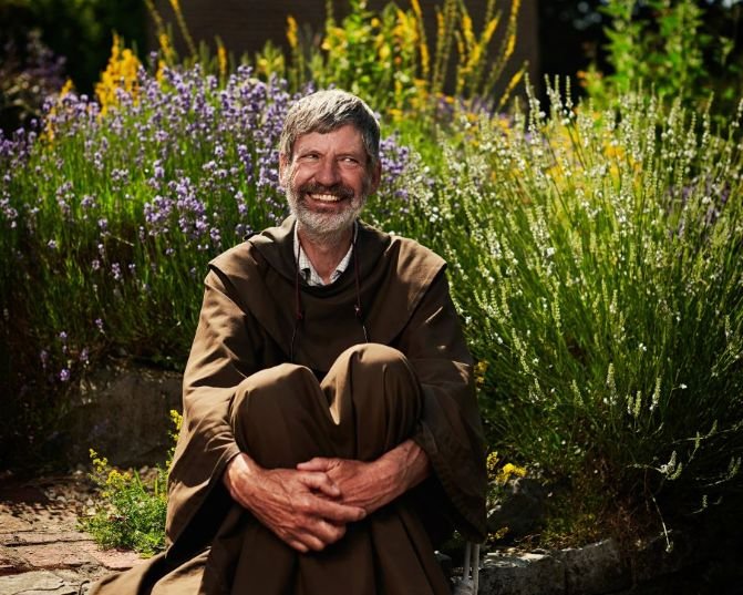 A person with grey hair and a beard, dressed in a brown robe, sits peacefully in a lush garden filled with vibrant purple and yellow flowers and greenery.