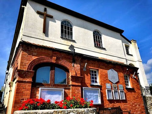 Exterior of Lyme Regis church with red brick and white plaster walls, arched windows, a wooden cross, and red flowers in planters.