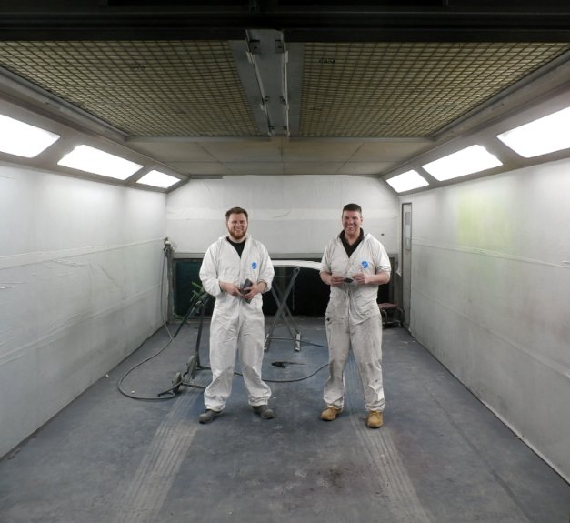 Two people in white coveralls inside a spray booth at Nuffield Accident Repair, holding small items with equipment and car parts in the background.